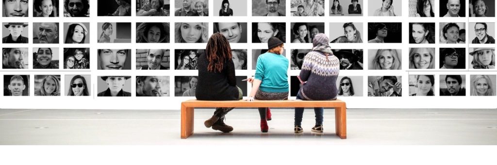 A wide shot of the backs of three people sitting in a gallery looking at a wall of photographs of faces