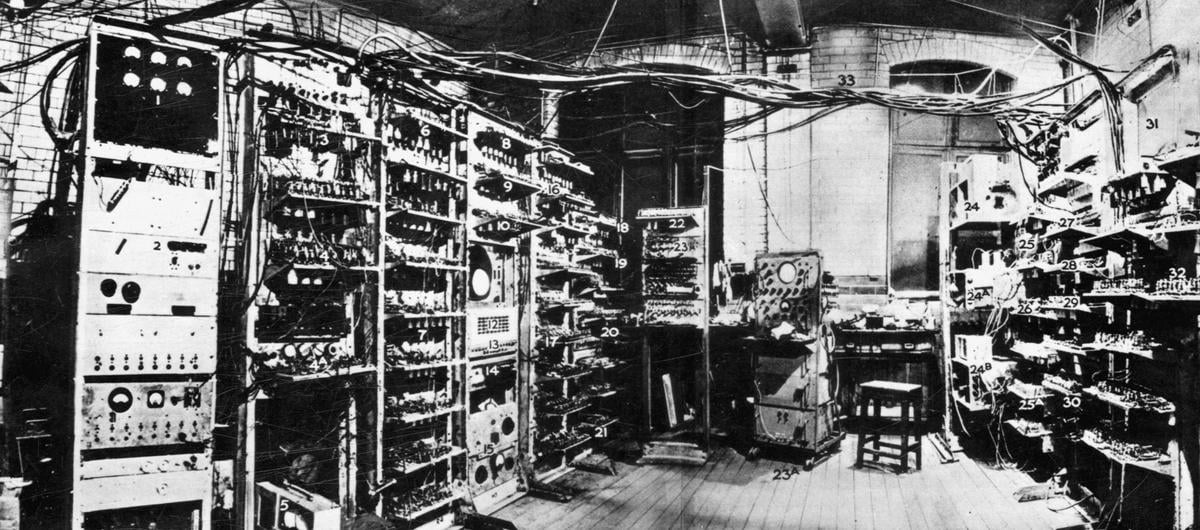 Black and white photograph of a computer room with equipment on racks and bundled wires across the ceiling.