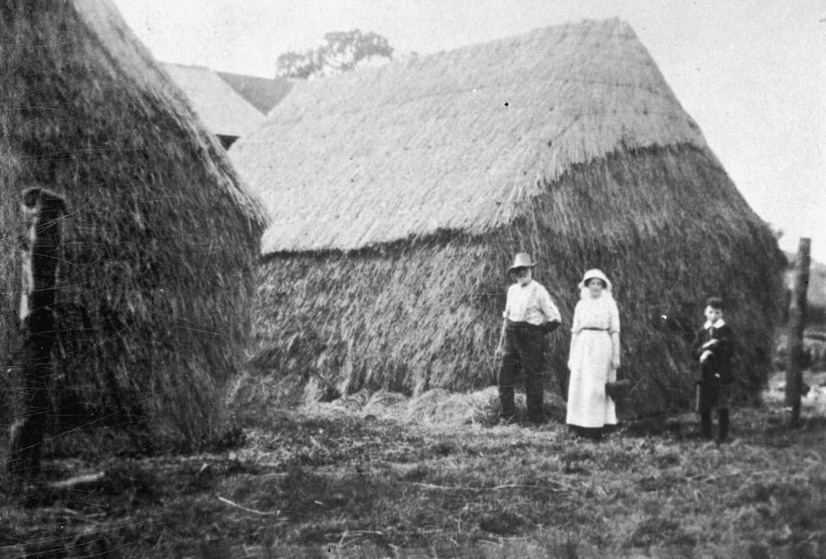Black and white photograph of a large stack of straw. Three figures stand in front of it.